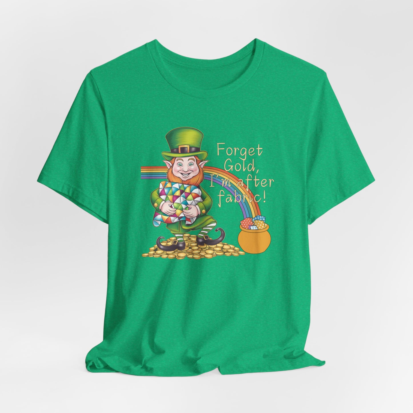 A Heather Kelly short-sleeve t-shirt featuring a cheerful leprechaun holding a quilt, sitting on gold coins, with a rainbow leading to a fabric-filled pot and the phrase 'Forget Gold, I’m After Fabric!' Perfect for quilters celebrating St. Patrick’s Day.