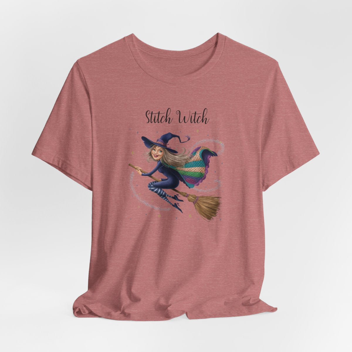 A Heather Mauve Halloween Quilting T-Shirt featuring a witch riding broomstick on t-shirt design. Colorful patchwork quilt cape with various patterns. Sparkling magic and stars. Text reads "Stitch Witch". Ideal for quilters who enjoy magical themes.