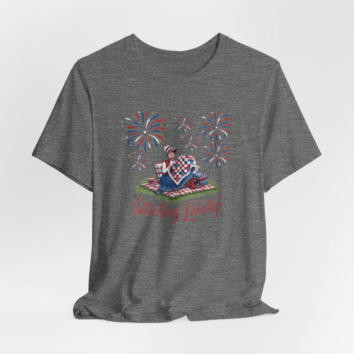 A Deep Heather T-shirt with illustrated scene of a colonial woman quilting a patriotic blanket, surrounded by fireworks. Text reads "Stitching Liberty" in a decorative font.