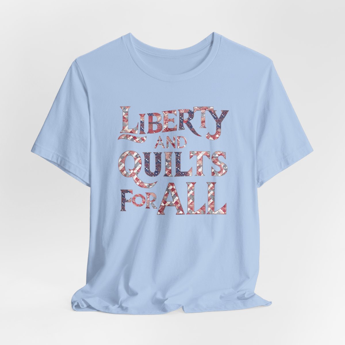 A Baby Blue T-shirt featuring text "LIBERTY AND QUILTS FOR ALL" in large decorative letters filled with various quilt patterns in red, white, and blue.