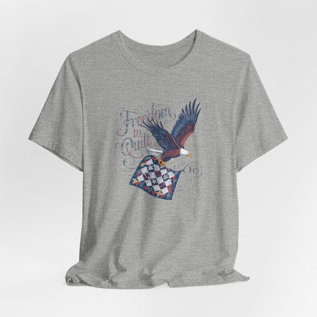 An Athletic Heather T-shirt with detailed illustration of a bald eagle in flight, carrying a patchwork quilt. Text reads "Freedom to Quilt" in an ornate, patriotic font.