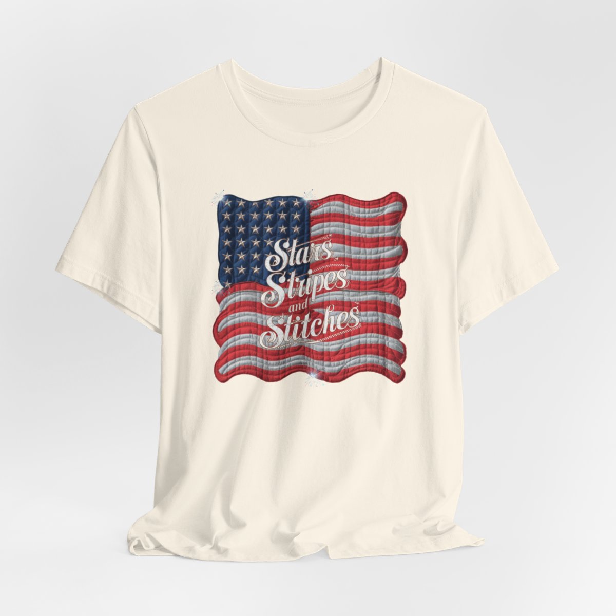 A Natural T-shirt featuring quilted American flag design with text "Stars Stripes and Stitches" overlaid in stylized, patriotic font. Fireworks accent the corners.