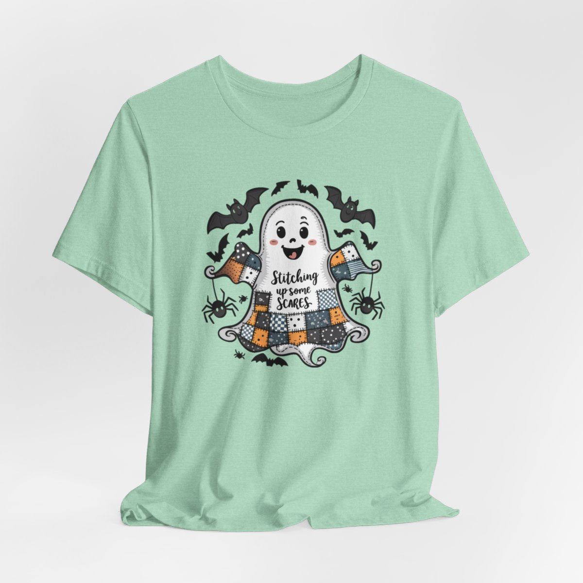 A Heather Mint Halloween Quilting T-Shirt featuring a smiling ghost with quilted patchwork body. Halloween-themed quilt patterns. Cute bats and spiders around ghost. Text reads "Stitching up some Scares". Ideal for quilters who enjoy Halloween themes.