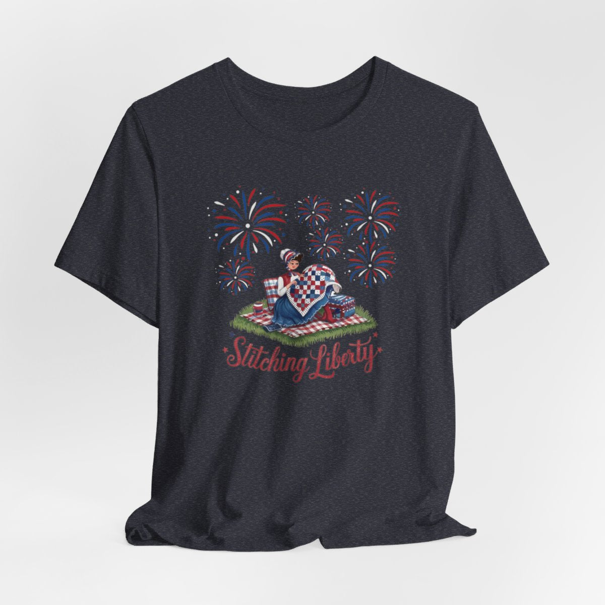 A Heather Navy T-shirt with illustrated scene of a colonial woman quilting a patriotic blanket, surrounded by fireworks. Text reads "Stitching Liberty" in a decorative font.