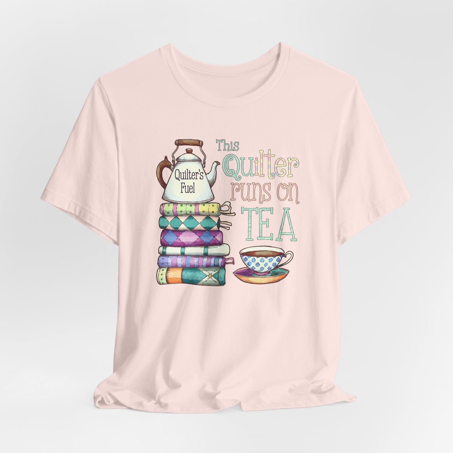 A Soft Pink t-shirt featuring a whimsical design of a stack of patchwork quilts with a teapot labeled 'Quilter's Fuel' and the phrase 'This Quilter Runs on Tea,' ideal for tea-loving quilters.