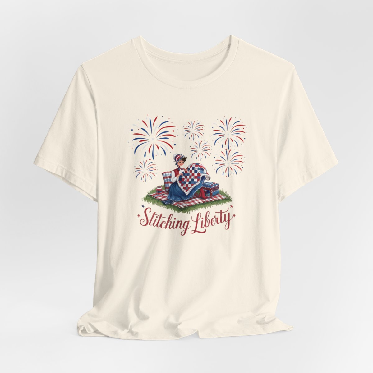 A Natural T-shirt with illustrated scene of a colonial woman quilting a patriotic blanket, surrounded by fireworks. Text reads "Stitching Liberty" in a decorative font.