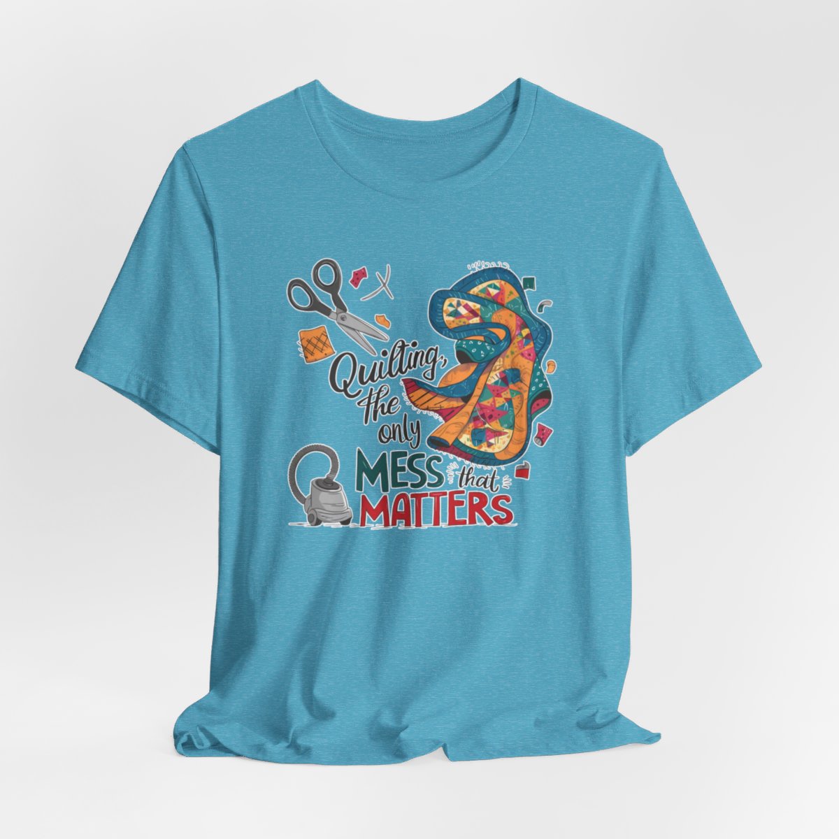 A Heather Aqua fun T-Shirt with the phrase Quilting, the Only Mess that Matters and illustrations of fabric scraps, scissors, and a vacuum cleaner. Colorful quilt patterns swirl around the text, creating a playful and relatable design for quilting enthusiasts.