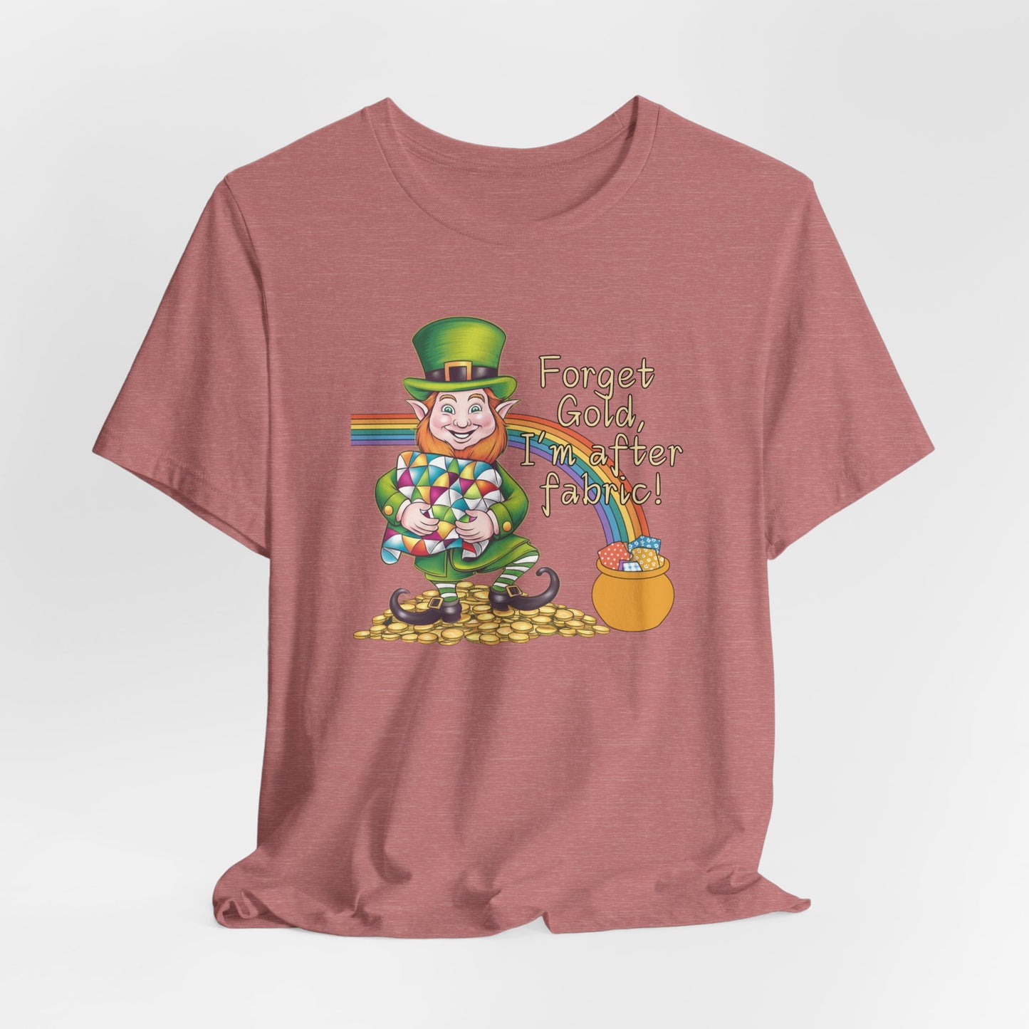 A Heather Mauve short-sleeve t-shirt featuring a cheerful leprechaun holding a quilt, sitting on gold coins, with a rainbow leading to a fabric-filled pot and the phrase 'Forget Gold, I’m After Fabric!' Perfect for quilters celebrating St. Patrick’s Day.