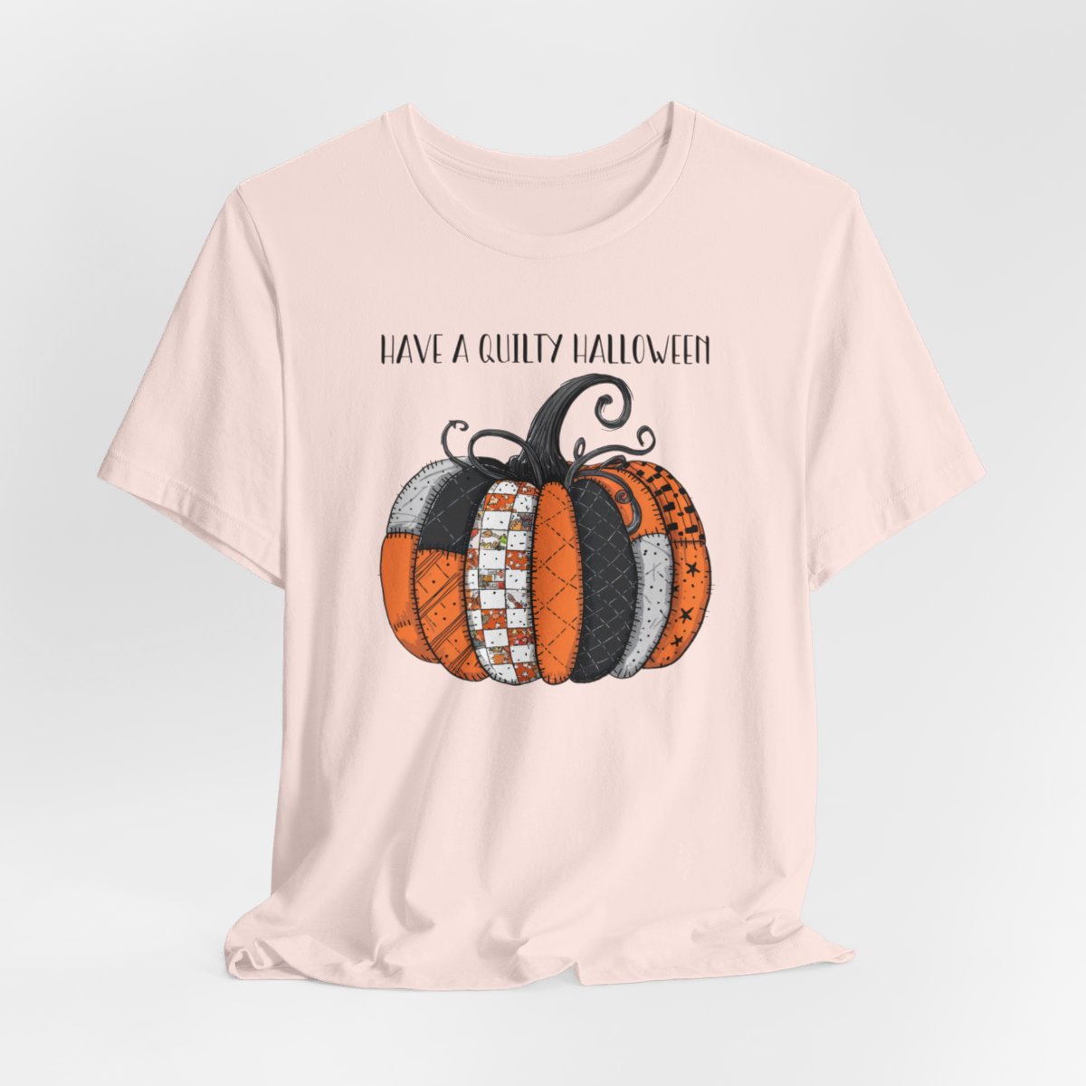 A Soft Pink Halloween Quilting T-Shirt featuring patchwork pumpkin with various quilt patterns. Mix of Halloween and traditional quilt motifs. Intricate stitching details. Text reads "Have a Quilty Halloween". Ideal for quilters who love Halloween.