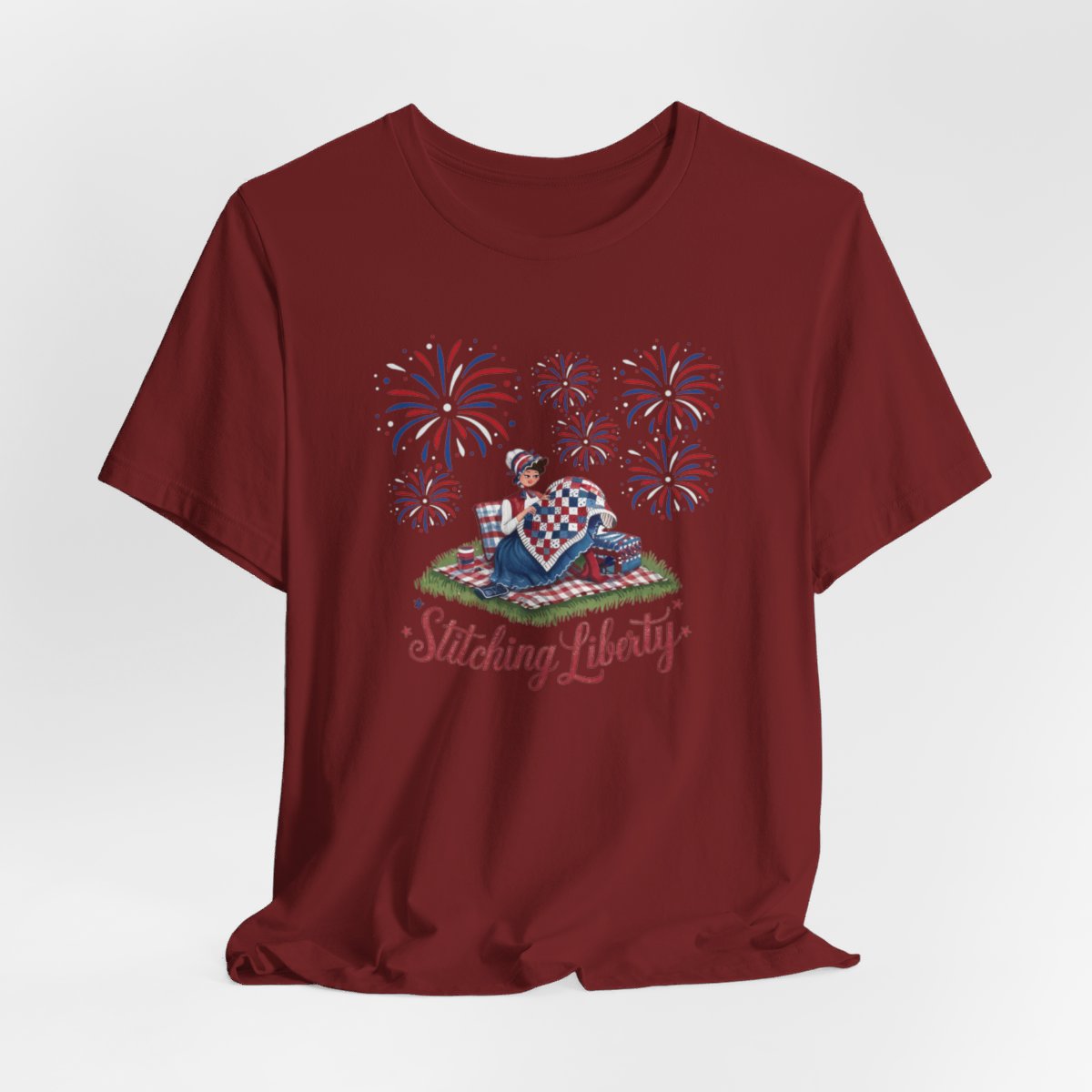 A Cardinal T-shirt with illustrated scene of a colonial woman quilting a patriotic blanket, surrounded by fireworks. Text reads "Stitching Liberty" in a decorative font.