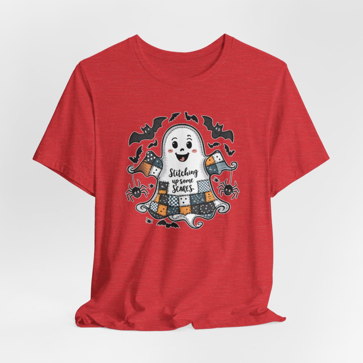 A Heather Red Halloween Quilting T-Shirt featuring a smiling ghost with quilted patchwork body. Halloween-themed quilt patterns. Cute bats and spiders around ghost. Text reads "Stitching up some Scares". Ideal for quilters who enjoy Halloween themes.