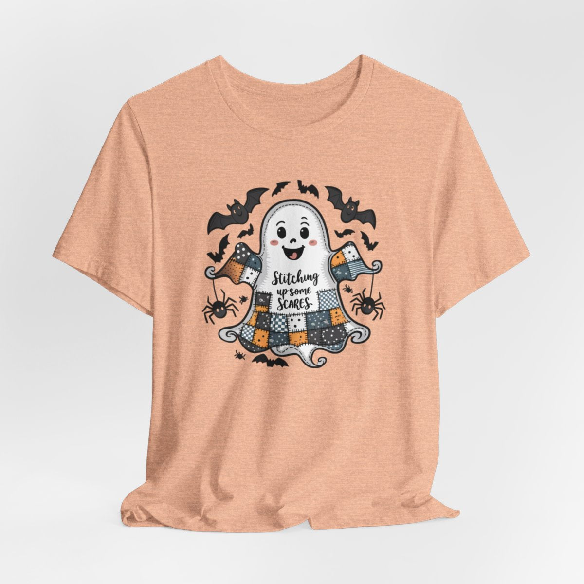 A Heather Peach Halloween Quilting T-Shirt featuring a smiling ghost with quilted patchwork body. Halloween-themed quilt patterns. Cute bats and spiders around ghost. Text reads "Stitching up some Scares". Ideal for quilters who enjoy Halloween themes.