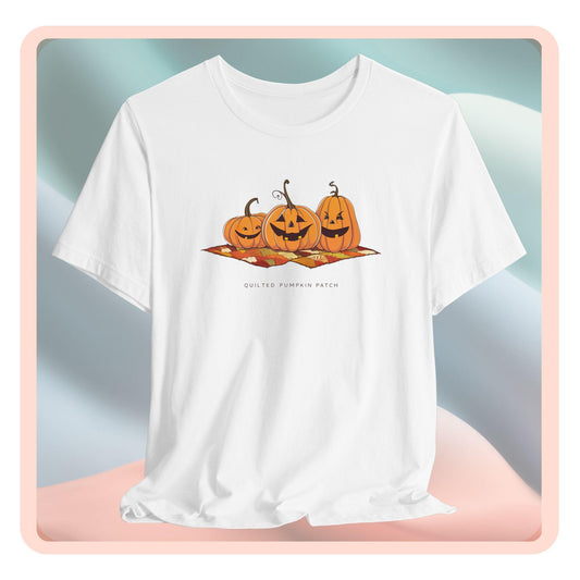 A T-shirt with three smiling jack-o'-lanterns sitting on a colorful autumn-themed quilt. The pumpkins have carved faces with various expressions, and the quilt beneath them features a patchwork of fall colors. Text below reads "QUILTED PUMPKIN PATCH".