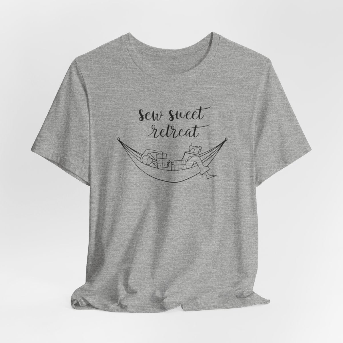 An Athletic Heather T-Shirt with a black and white illustration of a hammock filled with quilting supplies like fabric squares and a sewing machine. Text above reads "Sew Sweet Retreat" in a flowing script font.