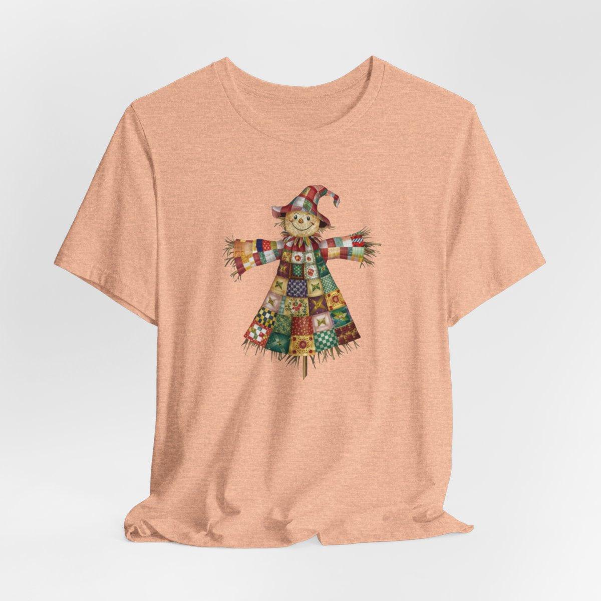 A Heather Peach thanksgiving  quilting t-shirt featuring a smiling scarecrow made entirely of colorful quilt patterns. Wearing a patchwork hat and clothes with various quilt designs. Straw details on hands and neck. Cheerful autumn-themed illustration celebrating quilting craft.