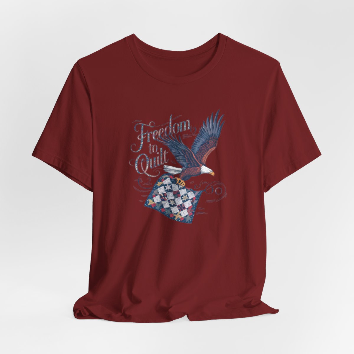 A Cardinal T-shirt with detailed illustration of a bald eagle in flight, carrying a patchwork quilt. Text reads "Freedom to Quilt" in an ornate, patriotic font.