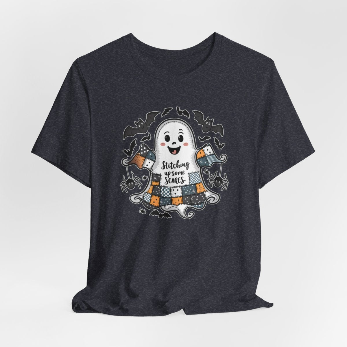 A Heather Navy Halloween Quilting T-Shirt featuring a smiling ghost with quilted patchwork body. Halloween-themed quilt patterns. Cute bats and spiders around ghost. Text reads "Stitching up some Scares". Ideal for quilters who enjoy Halloween themes.