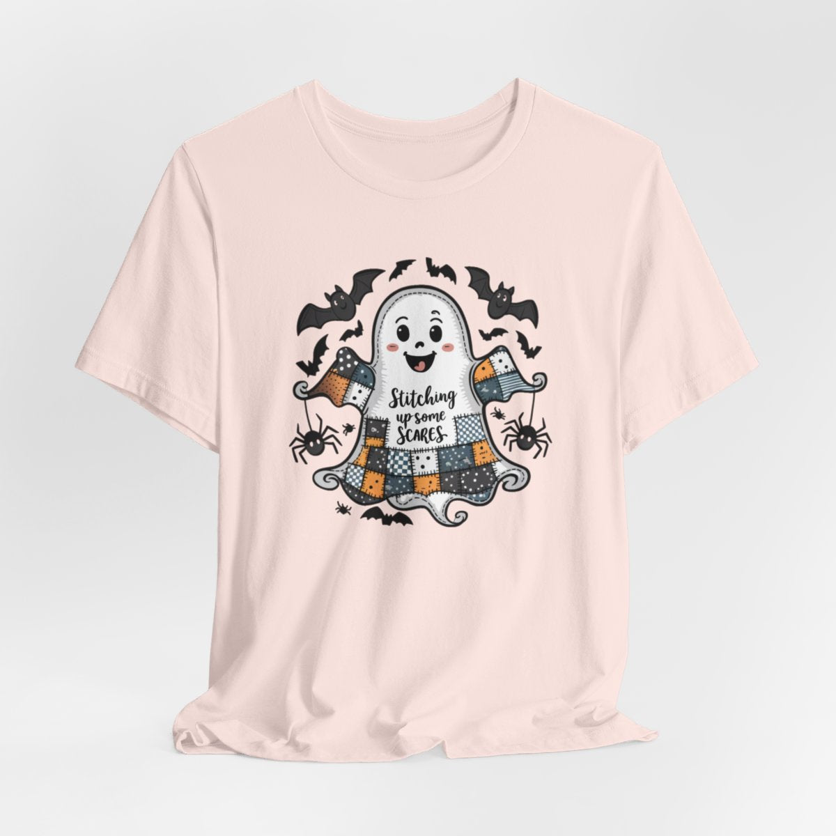 A Soft Pink Halloween Quilting T-Shirt featuring a smiling ghost with quilted patchwork body. Halloween-themed quilt patterns. Cute bats and spiders around ghost. Text reads "Stitching up some Scares". Ideal for quilters who enjoy Halloween themes.