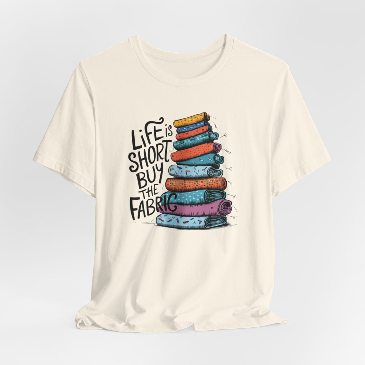 A Natural fun T-Shirt with the phrase Life is Short, Buy the Fabric with the label 'Life is Short, Buy the Fabric' next to a graphic of a fun pile of fabric