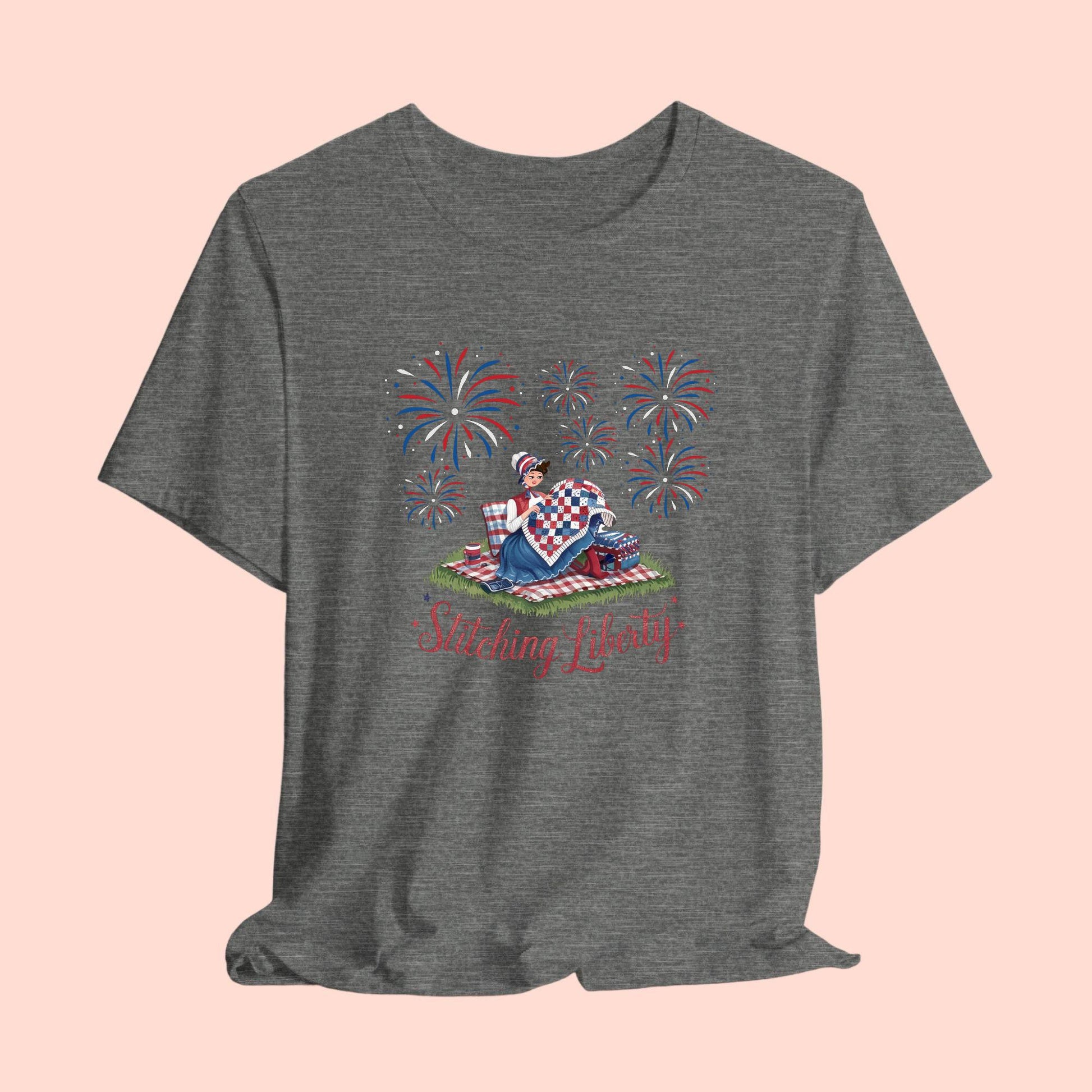 A T-shirt with illustrated scene of a colonial woman quilting a patriotic blanket, surrounded by fireworks. Text reads "Stitching Liberty" in a decorative font.