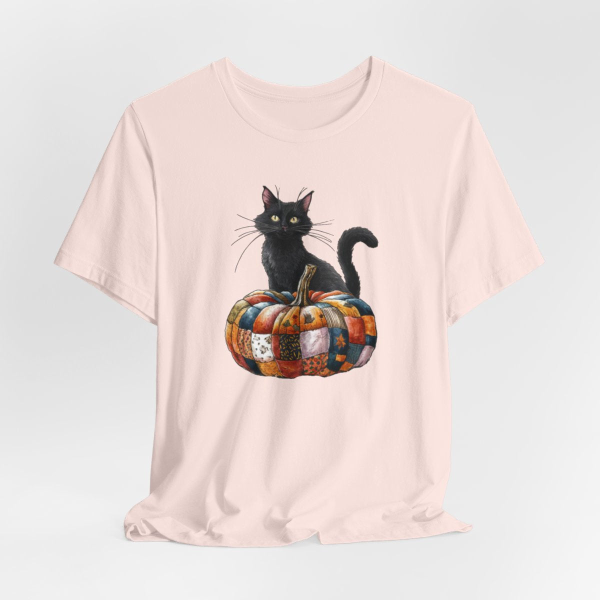 A Soft Pink Halloween Quilting T-Shirt featuring a black cat sitting on colorful patchwork quilt pumpkin. Cat has yellow eyes and long whiskers. Pumpkin features various quilt patterns in autumn colors. Halloween-themed t-shirt design for quilting enthusiasts.