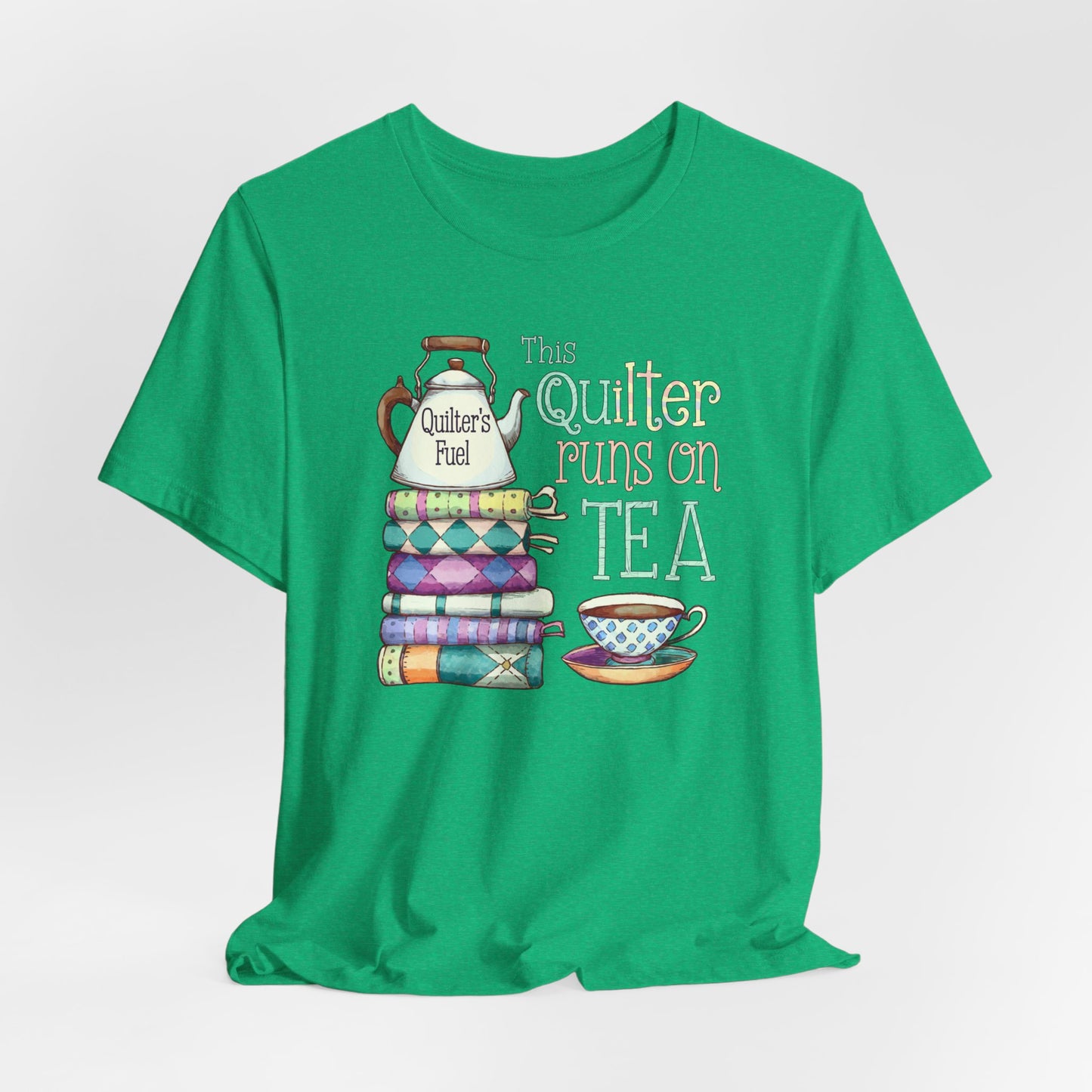 A Heather Kelly t-shirt featuring a whimsical design of a stack of patchwork quilts with a teapot labeled 'Quilter's Fuel' and the phrase 'This Quilter Runs on Tea,' ideal for tea-loving quilters.