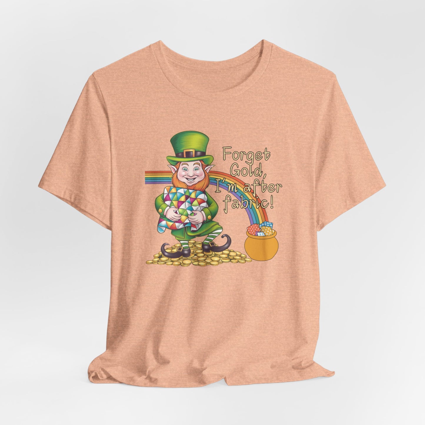 A Heather Peach short-sleeve t-shirt featuring a cheerful leprechaun holding a quilt, sitting on gold coins, with a rainbow leading to a fabric-filled pot and the phrase 'Forget Gold, I’m After Fabric!' Perfect for quilters celebrating St. Patrick’s Day.