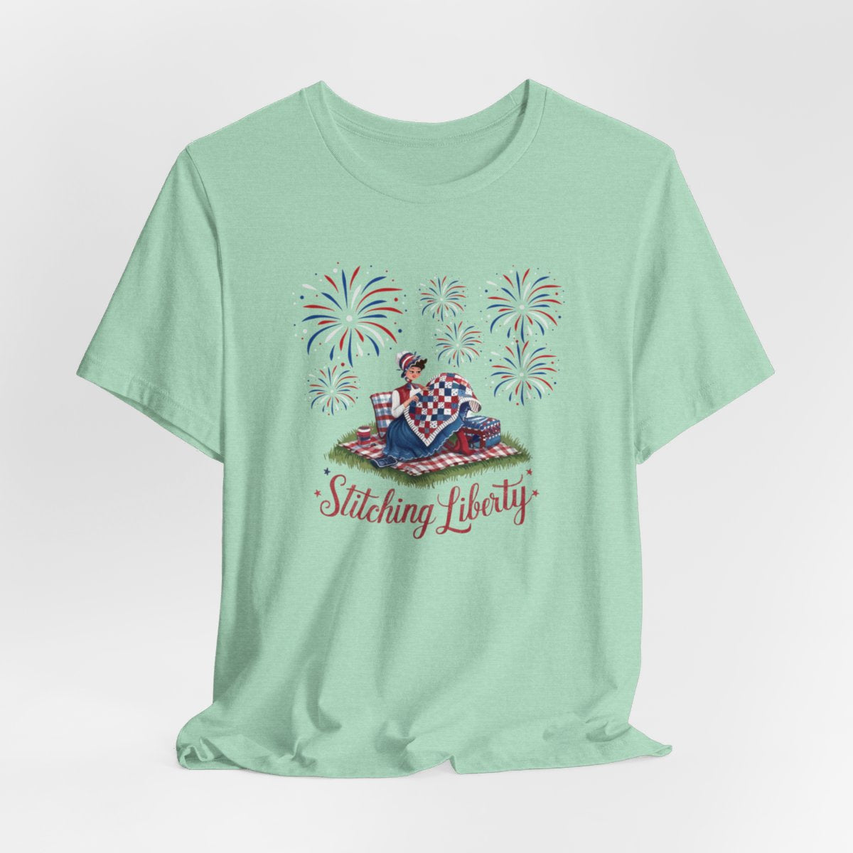 A Heather Mint T-shirt with illustrated scene of a colonial woman quilting a patriotic blanket, surrounded by fireworks. Text reads "Stitching Liberty" in a decorative font.