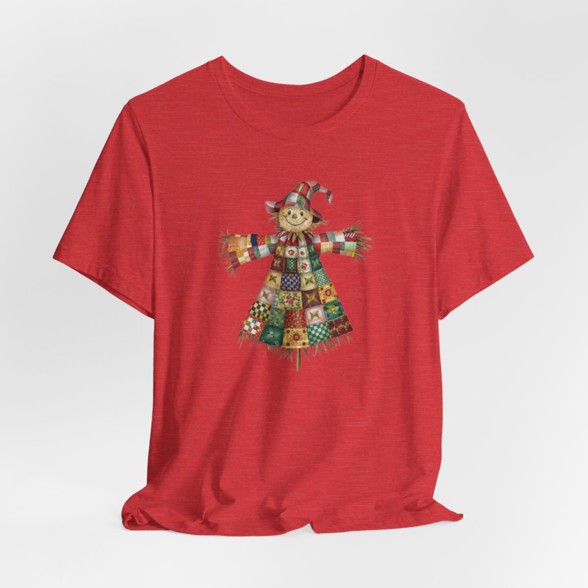 A Heather Red thanksgiving  quilting t-shirt featuring a smiling scarecrow made entirely of colorful quilt patterns. Wearing a patchwork hat and clothes with various quilt designs. Straw details on hands and neck. Cheerful autumn-themed illustration celebrating quilting craft.