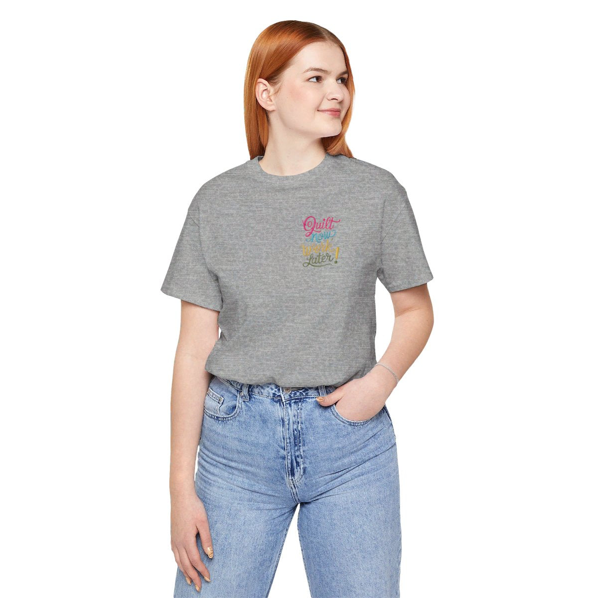 An Athletic Heather funny quilting T-shirt featuring bold, stylized text that reads "Quilt Now, Work Later" in various vibrant colors. The playful design captures the spirit of passionate quilters who prioritize their craft over other responsibilities.