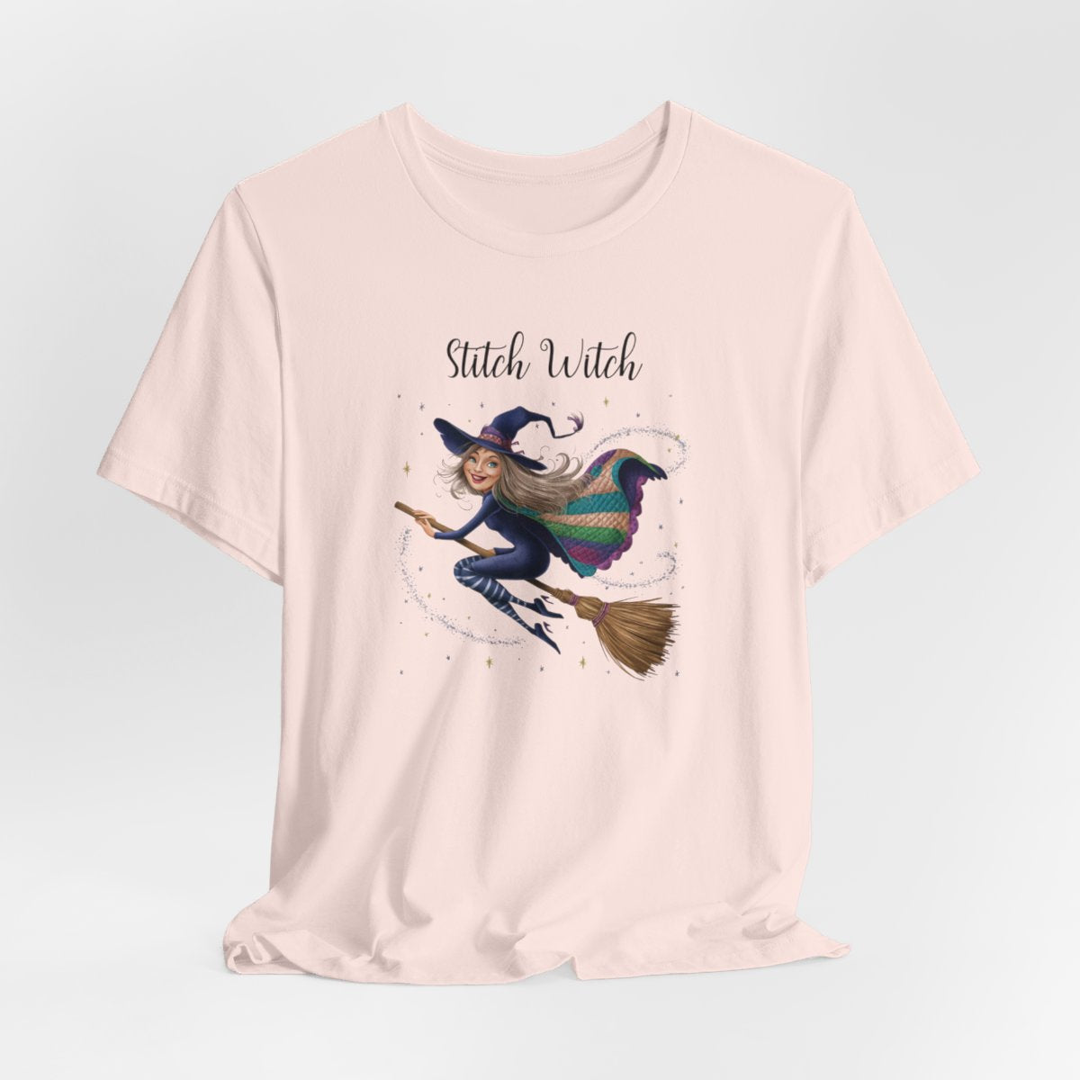 A Soft Pink Halloween Quilting T-Shirt featuring a witch riding broomstick on t-shirt design. Colorful patchwork quilt cape with various patterns. Sparkling magic and stars. Text reads "Stitch Witch". Ideal for quilters who enjoy magical themes.