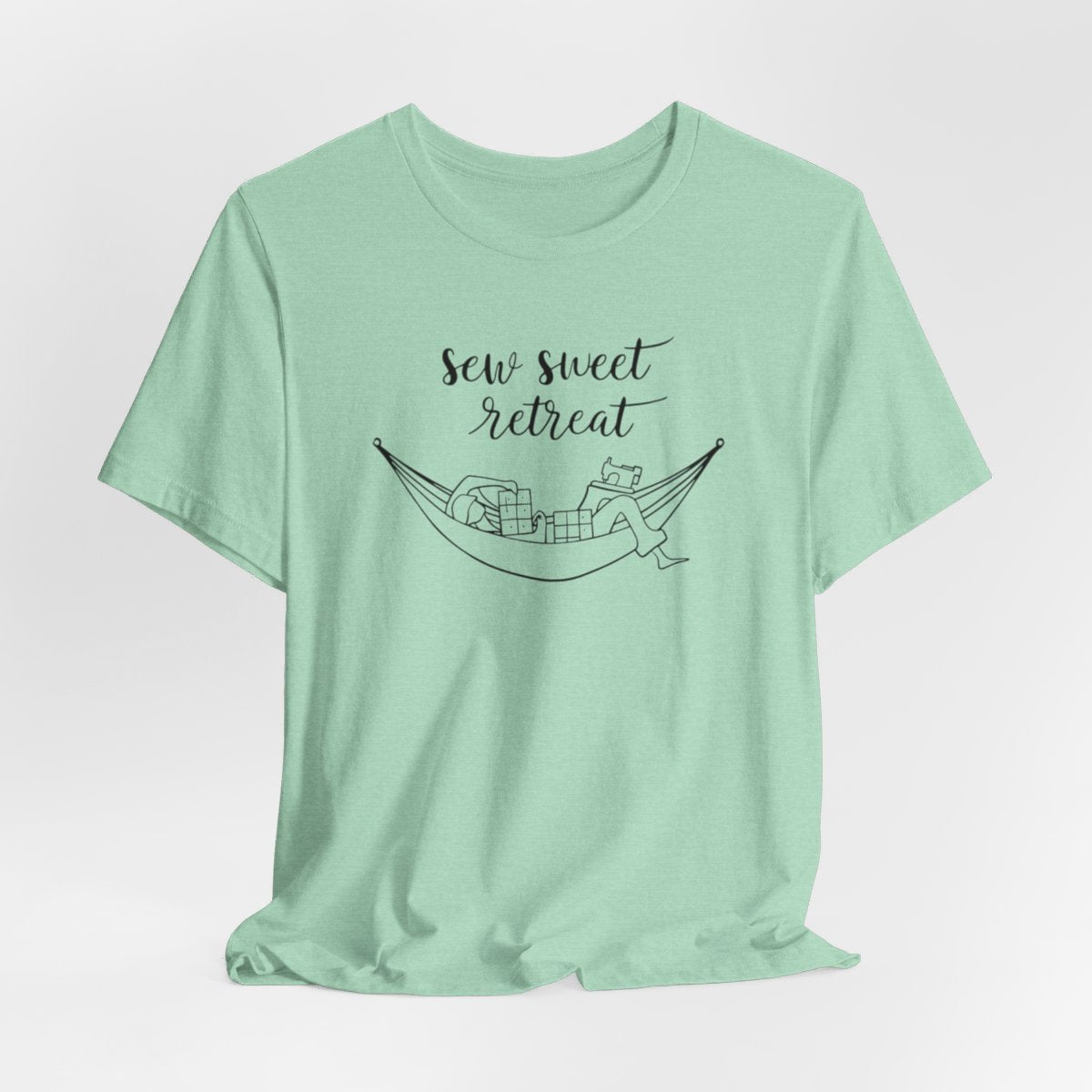 A Heather Mint T-Shirt with a black and white illustration of a hammock filled with quilting supplies like fabric squares and a sewing machine. Text above reads "Sew Sweet Retreat" in a flowing script font.