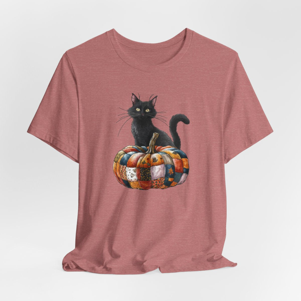 A Heather Mauve Halloween Quilting T-Shirt featuring a black cat sitting on colorful patchwork quilt pumpkin. Cat has yellow eyes and long whiskers. Pumpkin features various quilt patterns in autumn colors. Halloween-themed t-shirt design for quilting enthusiasts.