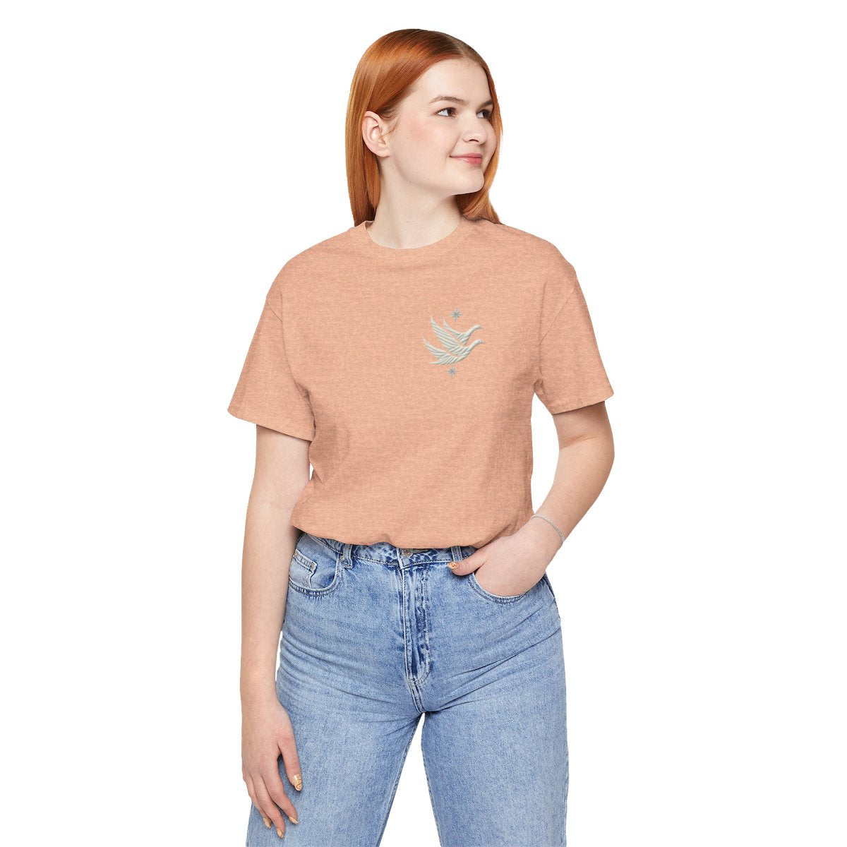 A Heather Peach T-shirt featuring two stylized white geese in flight, with outstretched wings. The geese are composed of intricate geometric patterns resembling quilt stitching. Two eight-pointed stars frame the scene, one above and one below the geese, adding a celestial element to the peaceful composition.