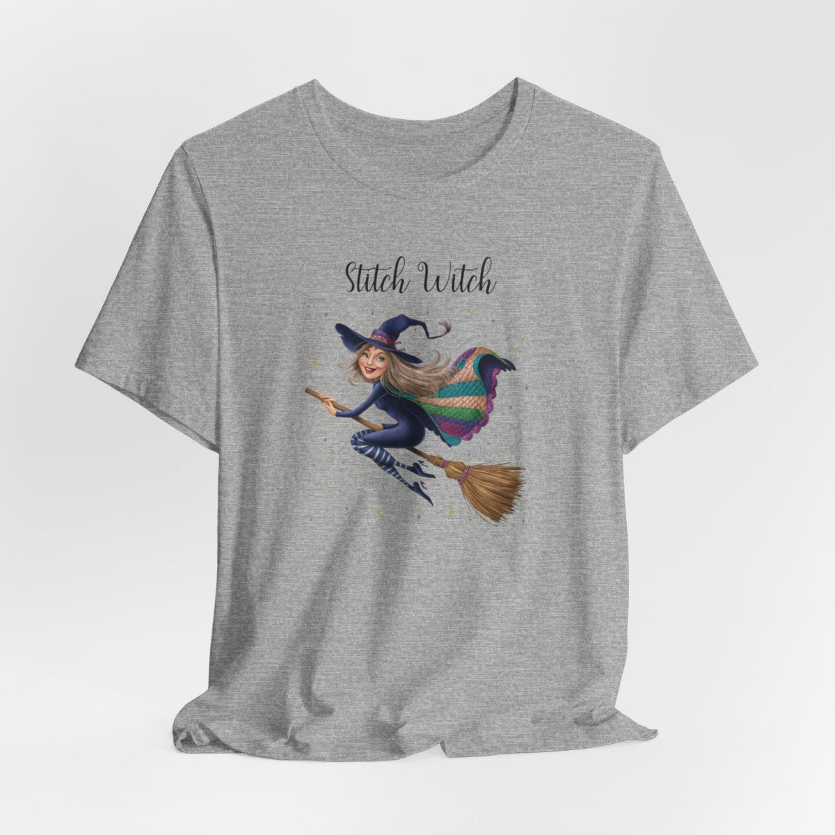 An Athletic Heather Halloween Quilting T-Shirt featuring a witch riding broomstick on t-shirt design. Colorful patchwork quilt cape with various patterns. Sparkling magic and stars. Text reads "Stitch Witch". Ideal for quilters who enjoy magical themes.