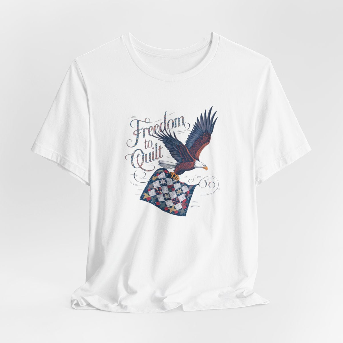 A White T-shirt with detailed illustration of a bald eagle in flight, carrying a patchwork quilt. Text reads "Freedom to Quilt" in an ornate, patriotic font.