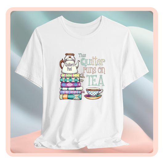 A t-shirt featuring a whimsical design of a stack of patchwork quilts with a teapot labeled 'Quilter's Fuel' and the phrase 'This Quilter Runs on Tea,' ideal for tea-loving quilters.