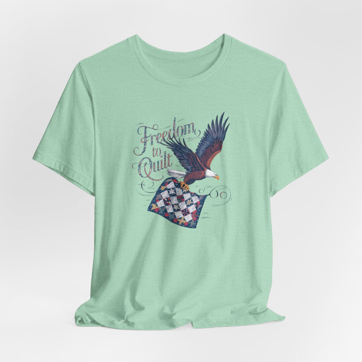 A Heather Mint T-shirt with detailed illustration of a bald eagle in flight, carrying a patchwork quilt. Text reads "Freedom to Quilt" in an ornate, patriotic font.