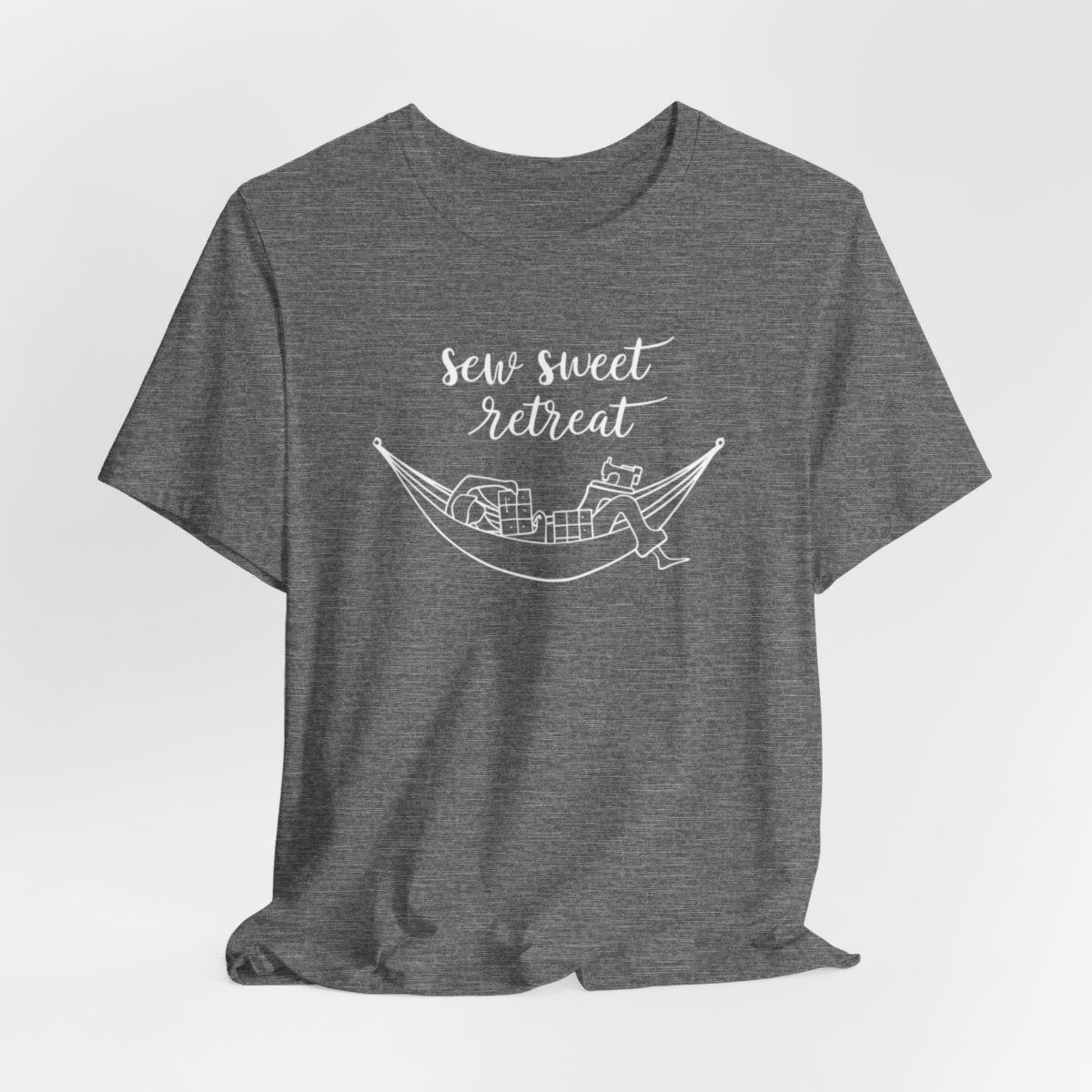 A Deep Heather T-Shirt with a black and white illustration of a hammock filled with quilting supplies like fabric squares and a sewing machine. Text above reads "Sew Sweet Retreat" in a flowing script font.