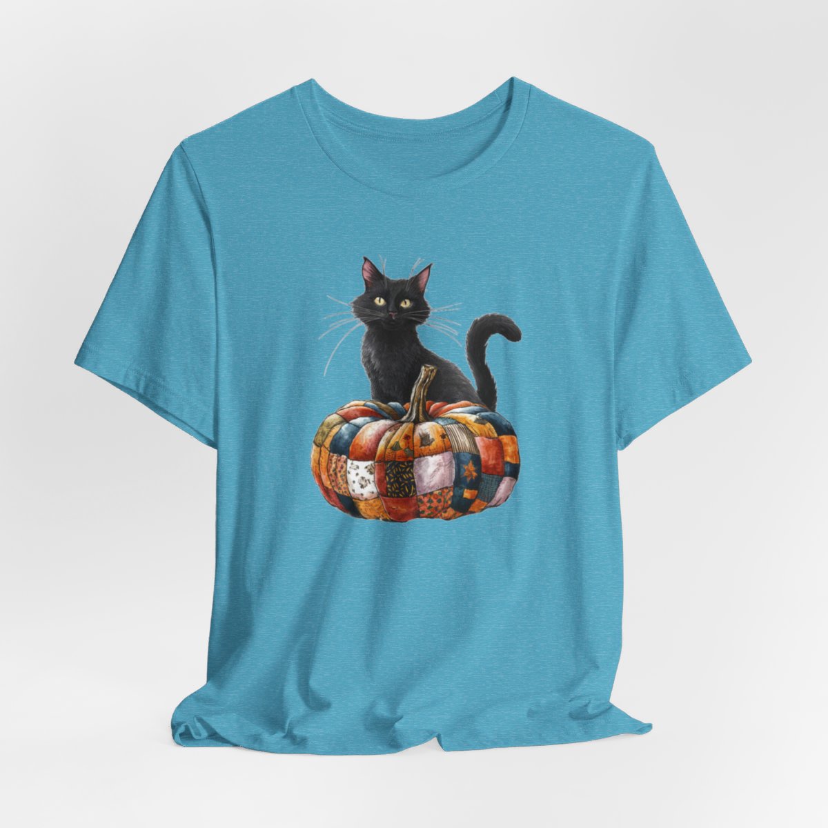 A Heather Aqua Halloween Quilting T-Shirt featuring a black cat sitting on colorful patchwork quilt pumpkin. Cat has yellow eyes and long whiskers. Pumpkin features various quilt patterns in autumn colors. Halloween-themed t-shirt design for quilting enthusiasts.