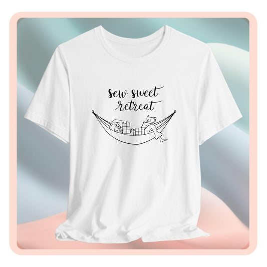 A T-Shirt with a black and white illustration of a hammock filled with quilting supplies like fabric squares and a sewing machine. Text above reads "Sew Sweet Retreat" in a flowing script font.