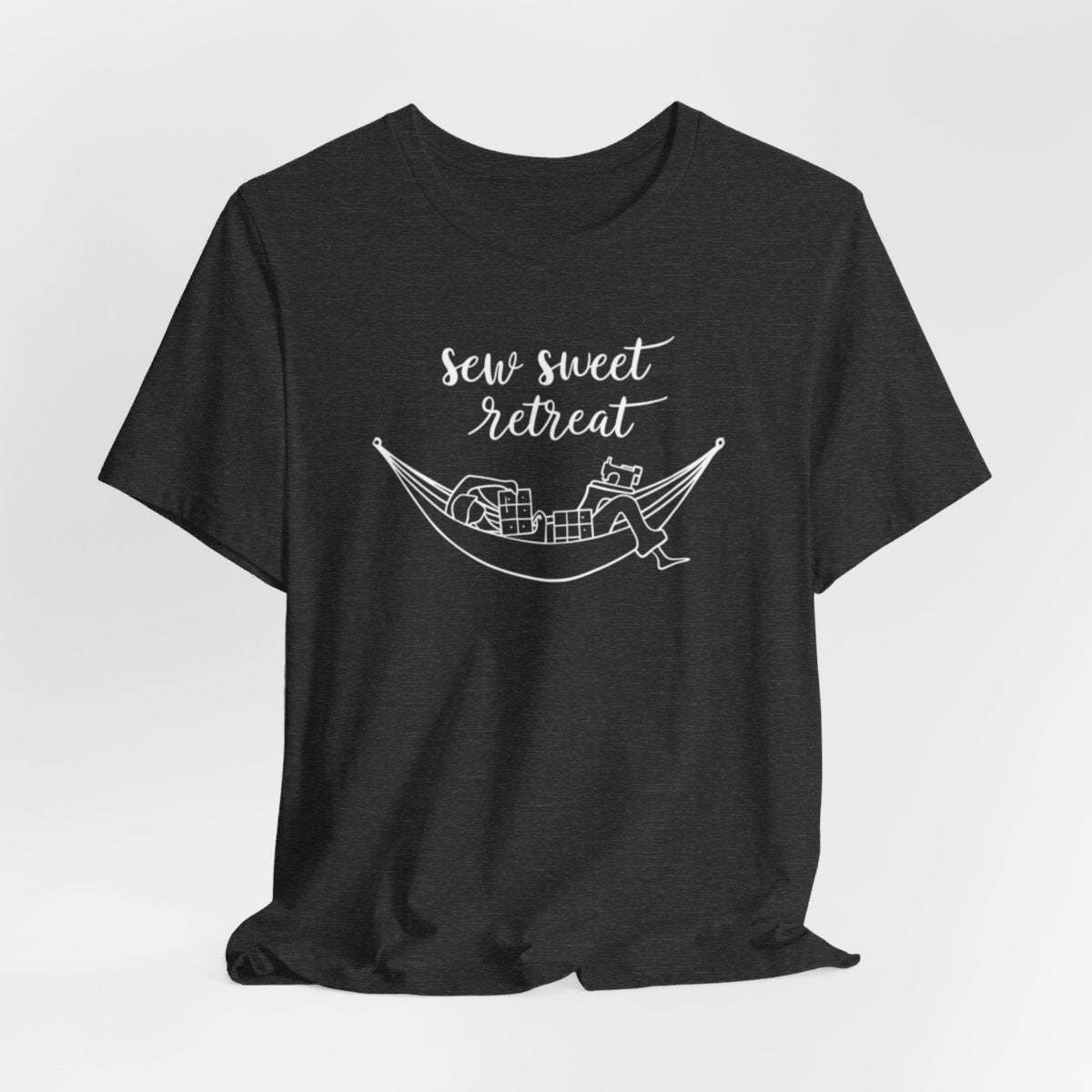 A Dark Grey Heather T-Shirt with a black and white illustration of a hammock filled with quilting supplies like fabric squares and a sewing machine. Text above reads "Sew Sweet Retreat" in a flowing script font.