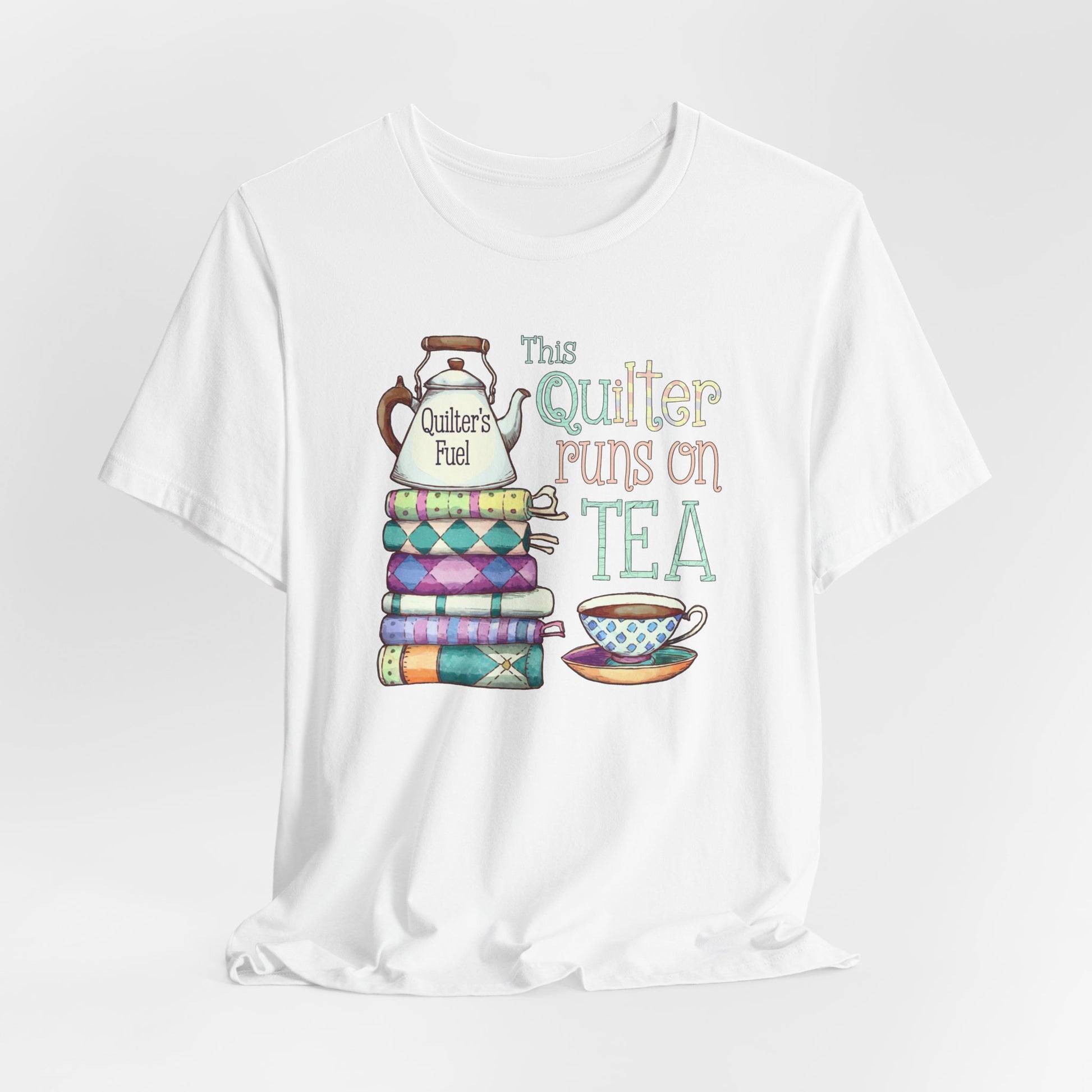 A White t-shirt featuring a whimsical design of a stack of patchwork quilts with a teapot labeled 'Quilter's Fuel' and the phrase 'This Quilter Runs on Tea,' ideal for tea-loving quilters.