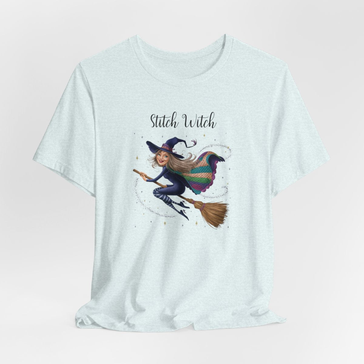 A Heather Ice Blue Halloween Quilting T-Shirt featuring a witch riding broomstick on t-shirt design. Colorful patchwork quilt cape with various patterns. Sparkling magic and stars. Text reads "Stitch Witch". Ideal for quilters who enjoy magical themes.