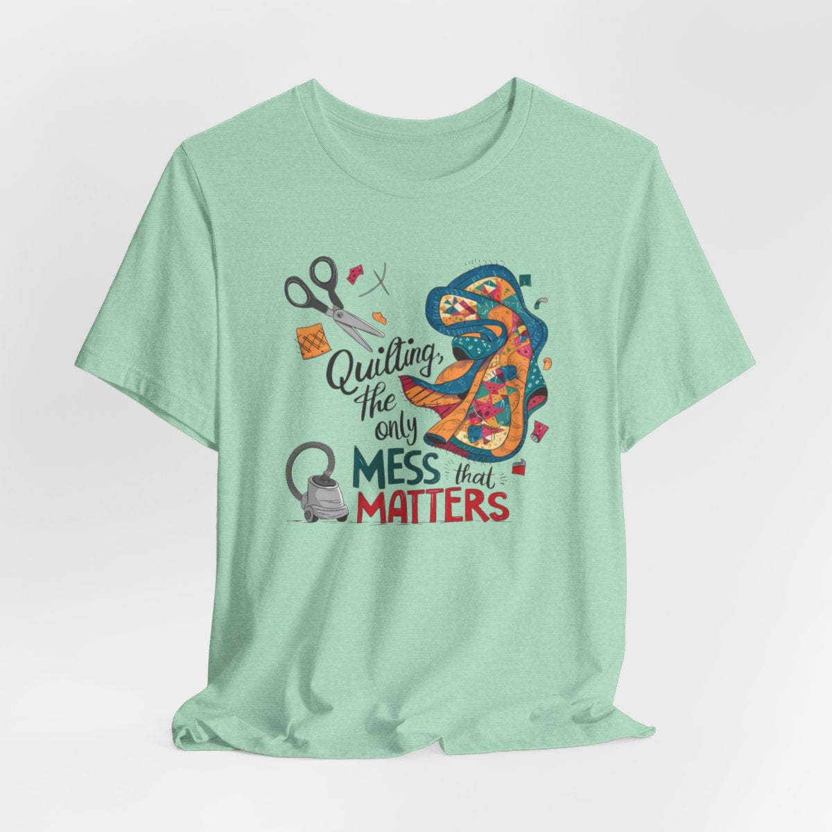 A Heather Mint fun T-Shirt with the phrase Quilting, the Only Mess that Matters and illustrations of fabric scraps, scissors, and a vacuum cleaner. Colorful quilt patterns swirl around the text, creating a playful and relatable design for quilting enthusiasts.