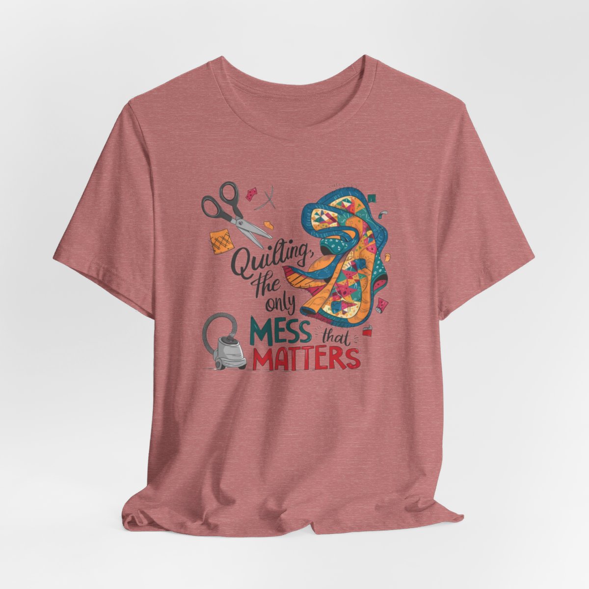 A Heather Mauve fun T-Shirt with the phrase Quilting, the Only Mess that Matters and illustrations of fabric scraps, scissors, and a vacuum cleaner. Colorful quilt patterns swirl around the text, creating a playful and relatable design for quilting enthusiasts.