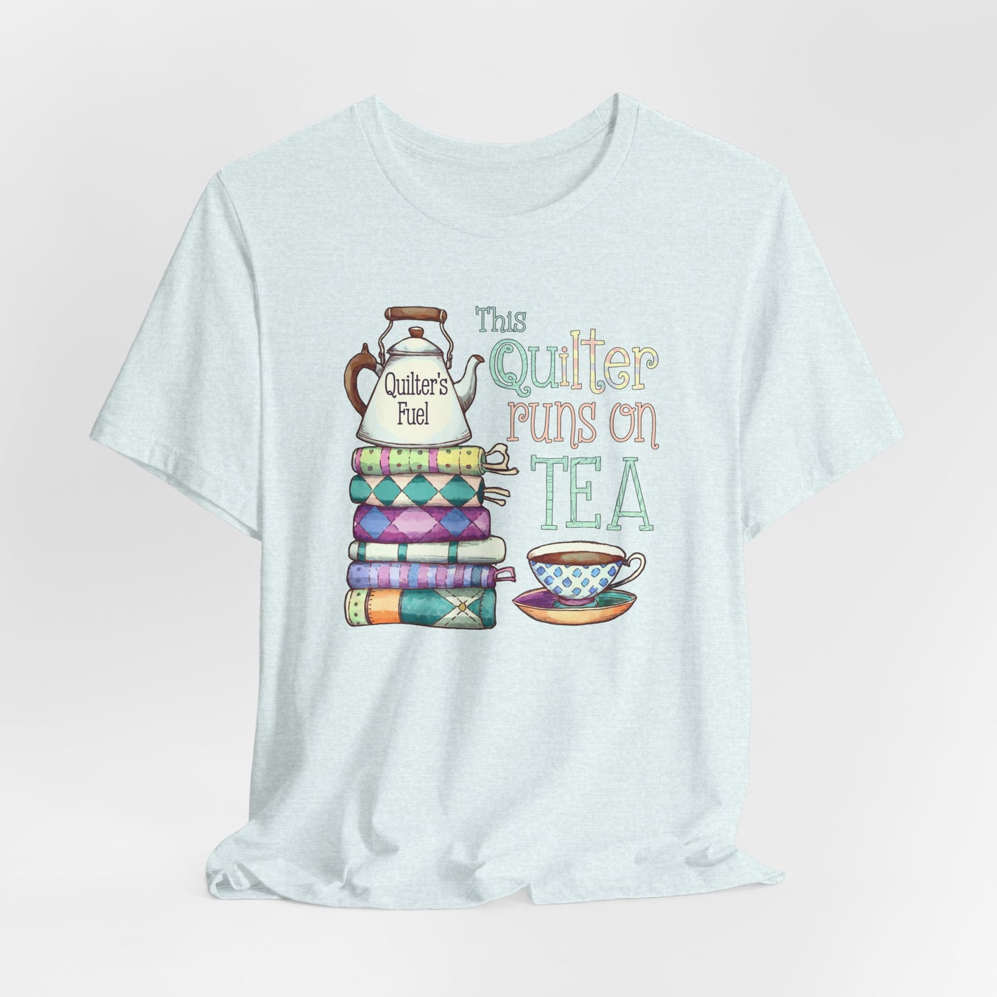 A Heather Ice Blue t-shirt featuring a whimsical design of a stack of patchwork quilts with a teapot labeled 'Quilter's Fuel' and the phrase 'This Quilter Runs on Tea,' ideal for tea-loving quilters.