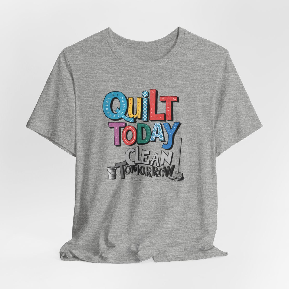 An Athletic Heather funny quilting T-Shirt with the phrase Quilt Today, Clean Tomorrow in a bold and cheerful font with a broom off to the side