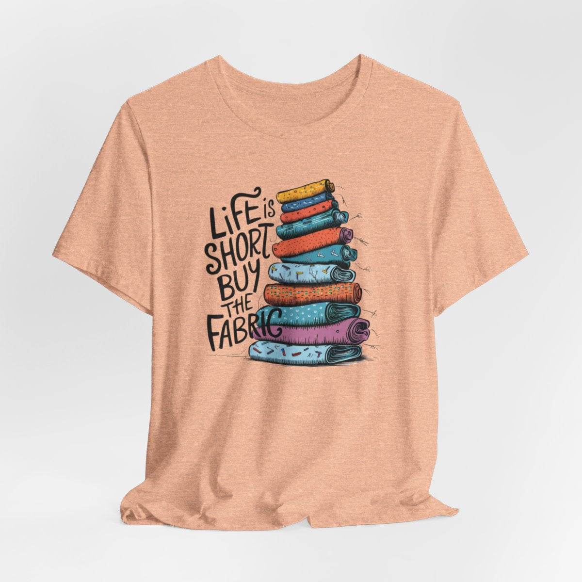 A Heather Peach fun T-Shirt with the phrase Life is Short, Buy the Fabric with the label 'Life is Short, Buy the Fabric' next to a graphic of a fun pile of fabric