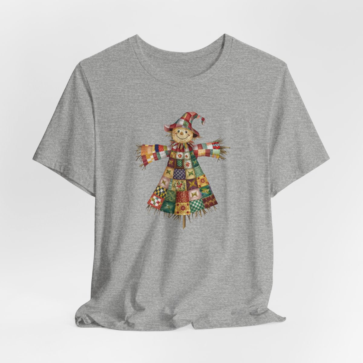 An Athletic Heather thanksgiving  quilting t-shirt featuring a smiling scarecrow made entirely of colorful quilt patterns. Wearing a patchwork hat and clothes with various quilt designs. Straw details on hands and neck. Cheerful autumn-themed illustration celebrating quilting craft.